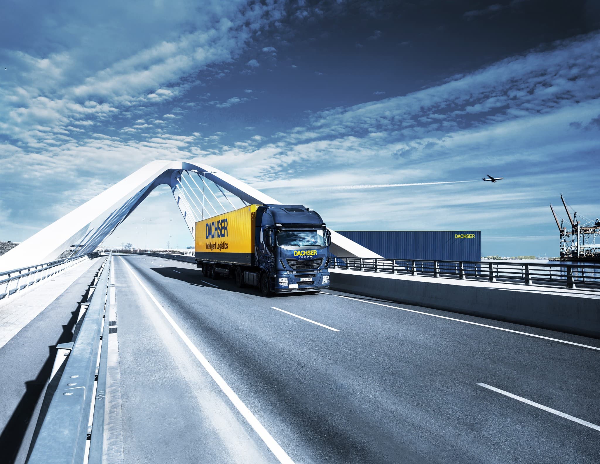 DACHSER logistics truck crossing a modern white bridge with a warehouse and airplane in the background under a blue sky.
