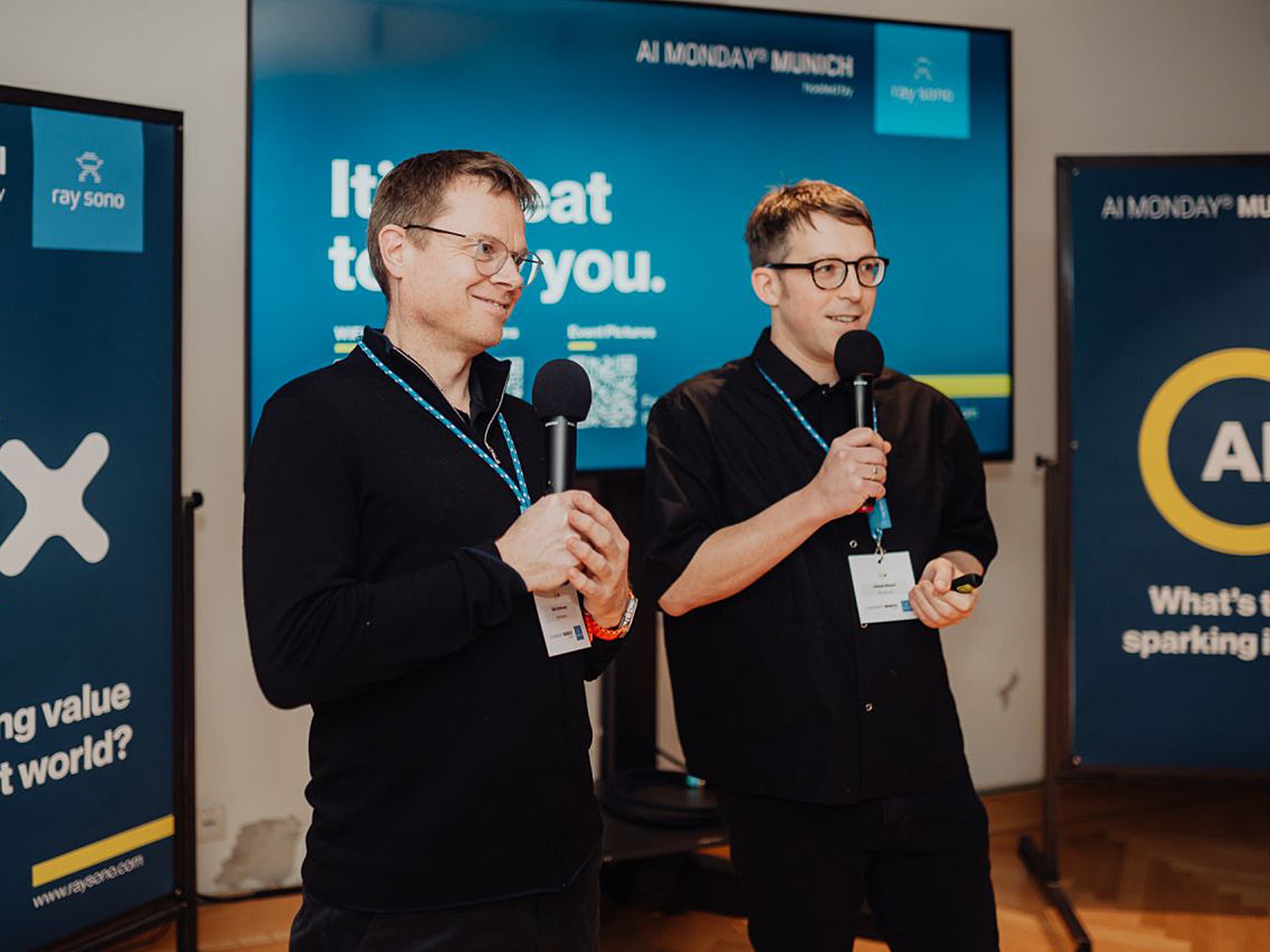 Two speakers with microphones standing in front of AI MONDAY® Munich event banners and a screen, wearing lanyards and name badges.