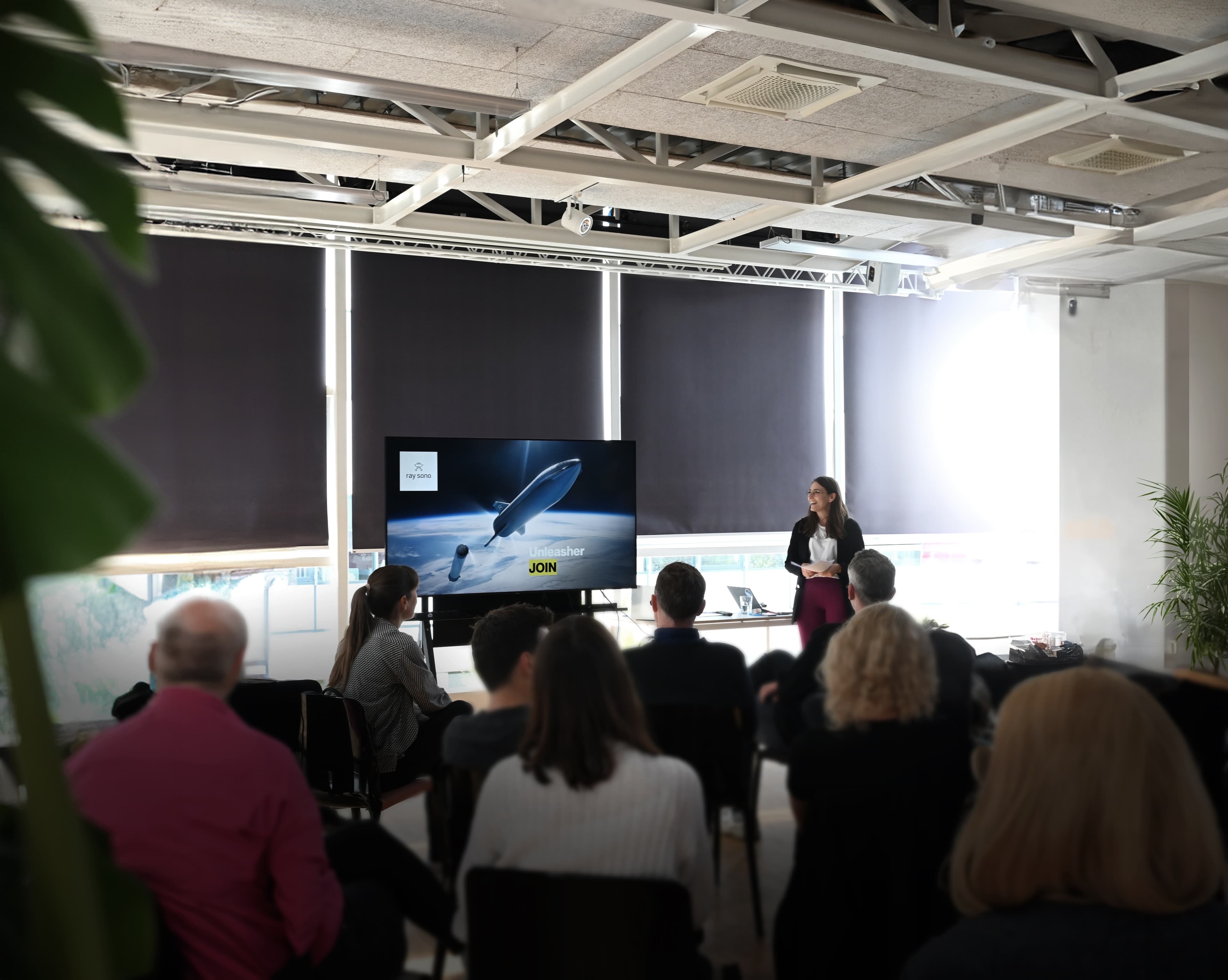 Woman giving a presentation to a seated audience, with a large screen showing a futuristic airship.