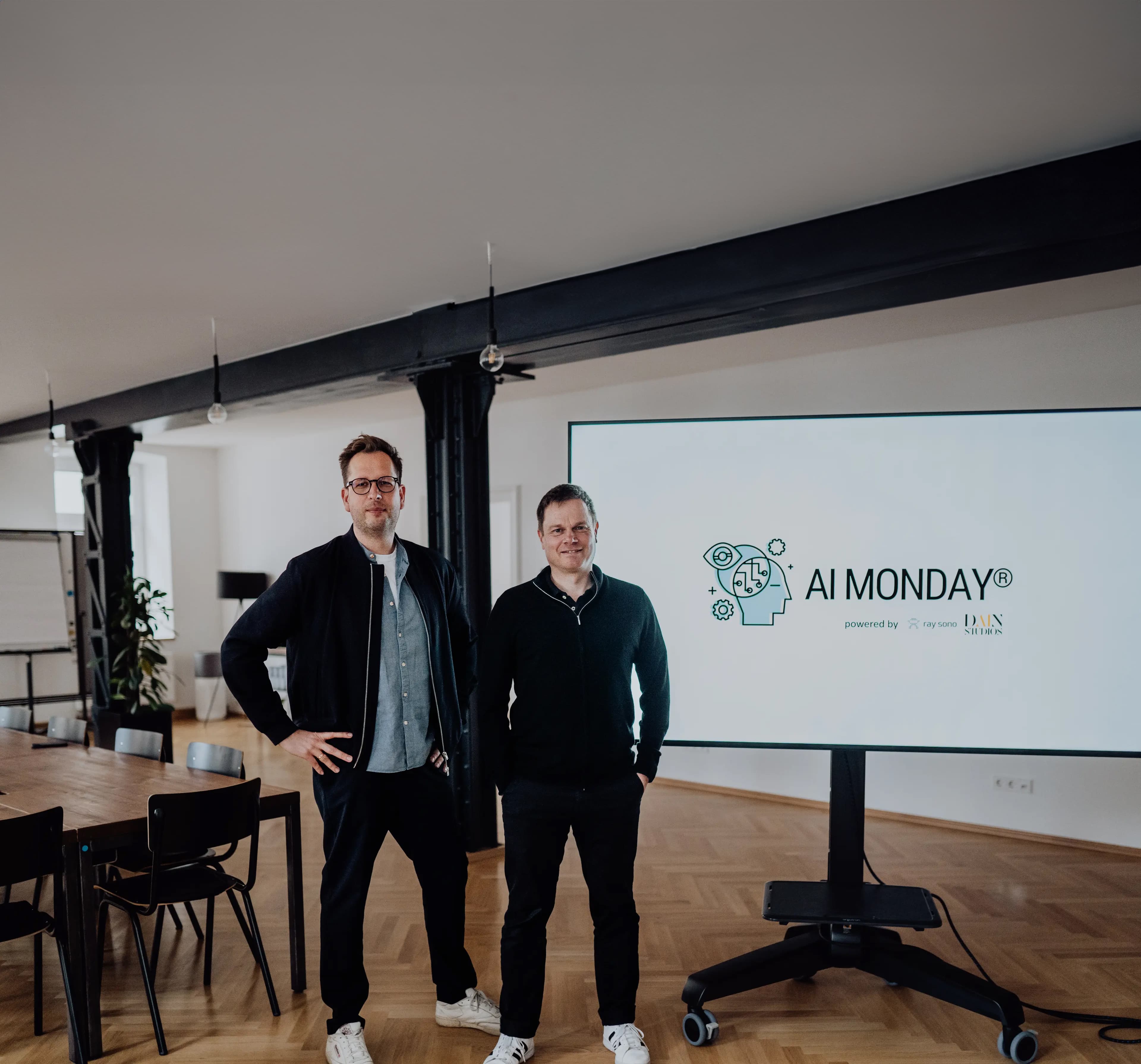 Two men standing next to a large screen displaying the logo and branding of AI MONDAY® in a modern meeting room.
