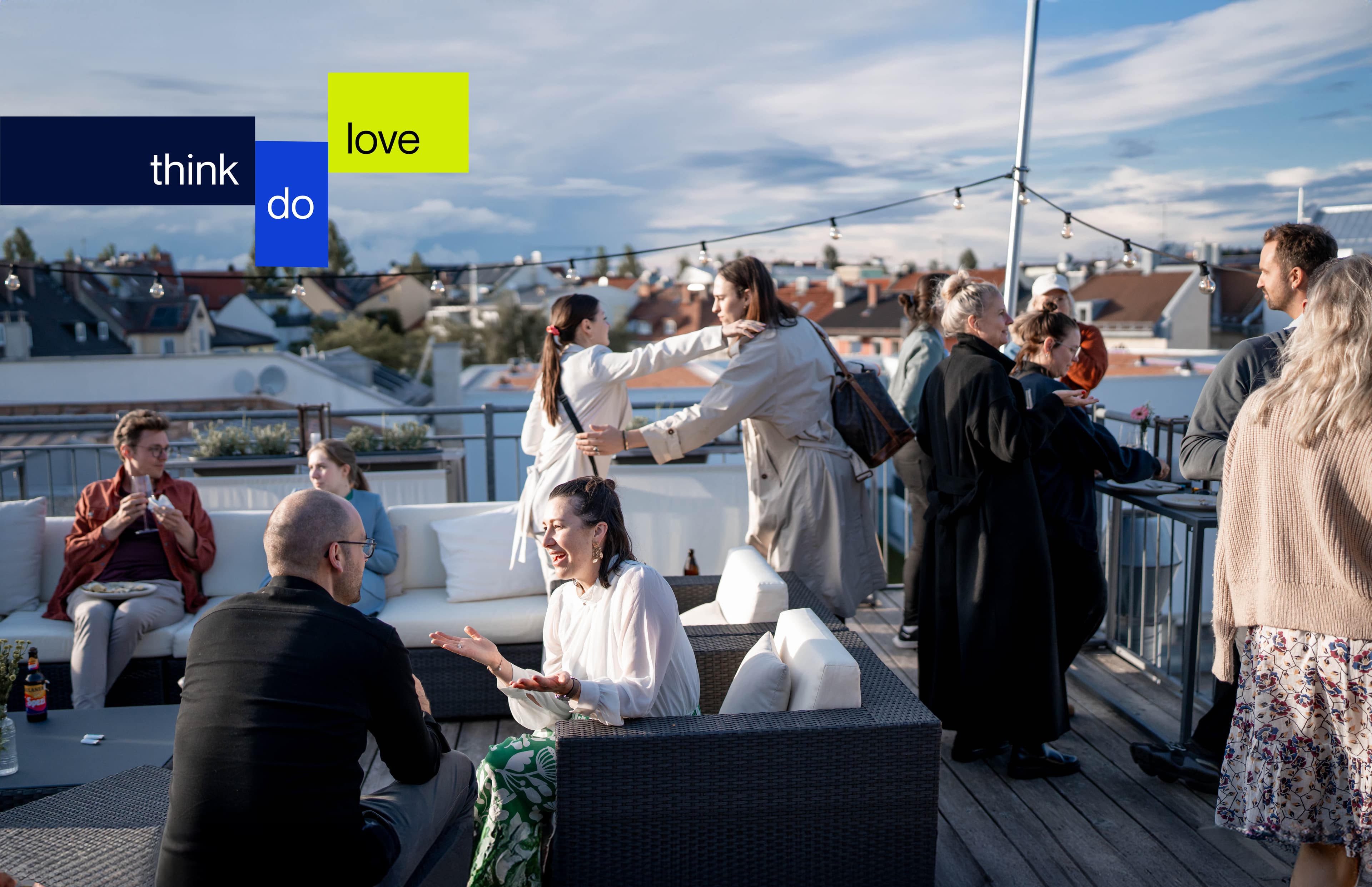 People socializing on a rooftop terrace with a view over city rooftops, under a partly cloudy sky, with the Ray Sono slogan think do love overlaid in bold colored blocks.