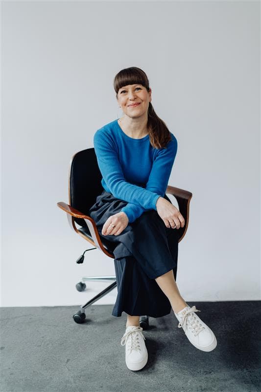 Woman in a blue sweater and white sneakers sitting casually on a swivel chair in front of a light background, smiling.