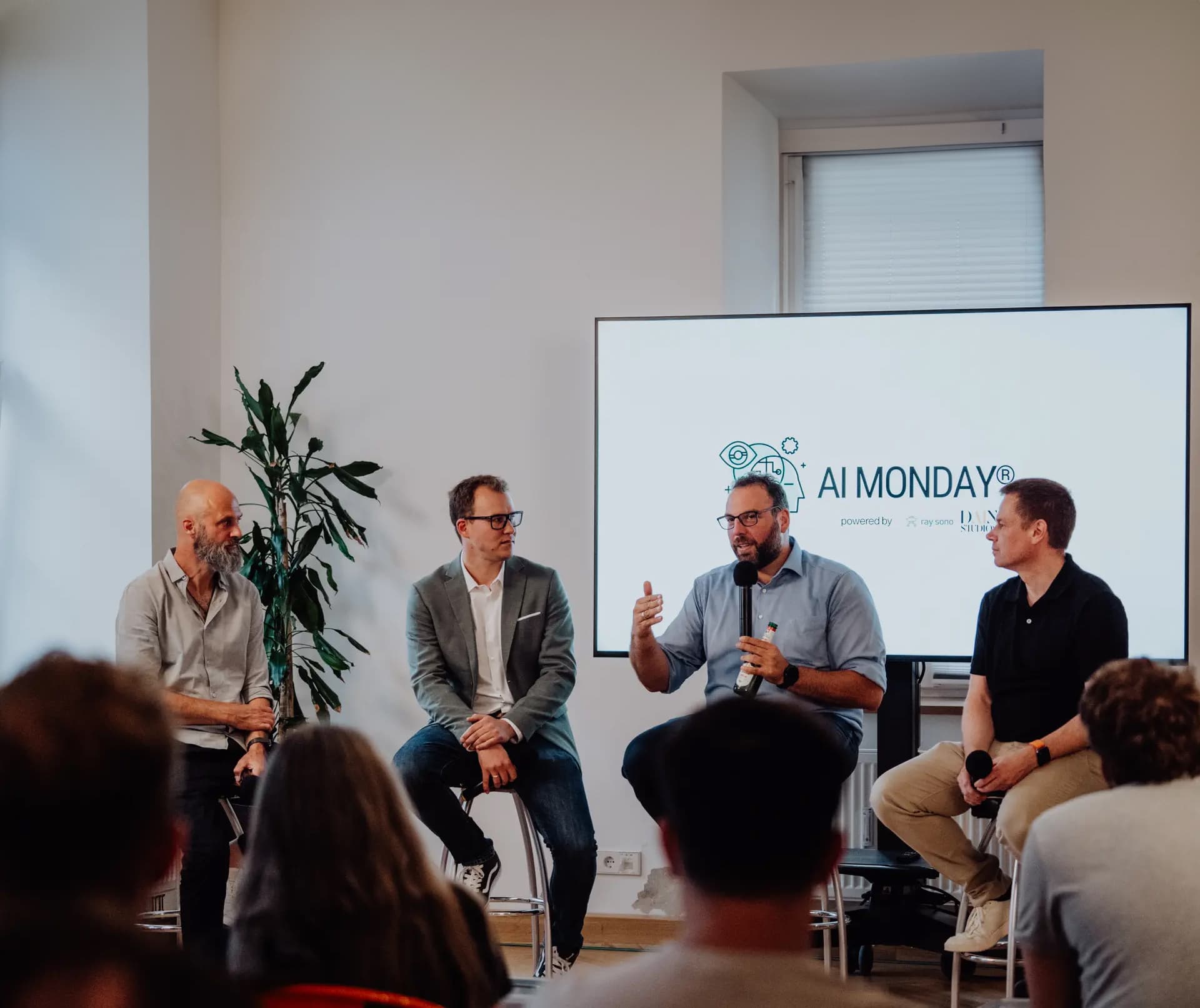 Four men in discussion on a panel at an AI MONDAY® event, seated in front of a screen with the event branding, while an audience listens.