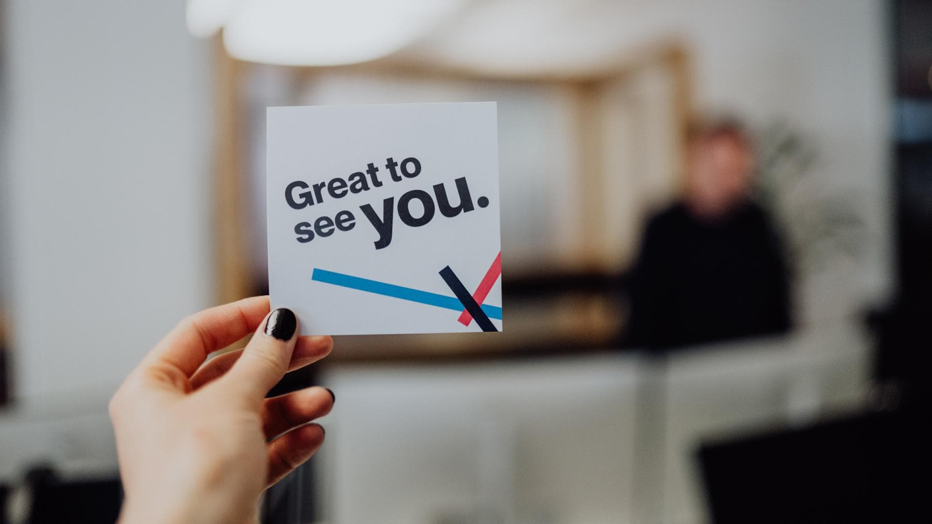 Hand hält eine gedruckte Karte mit der Aufschrift Great to see you in kräftiger Typografie vor dem unscharfen Hintergrund eines modernen Empfangsbereichs.