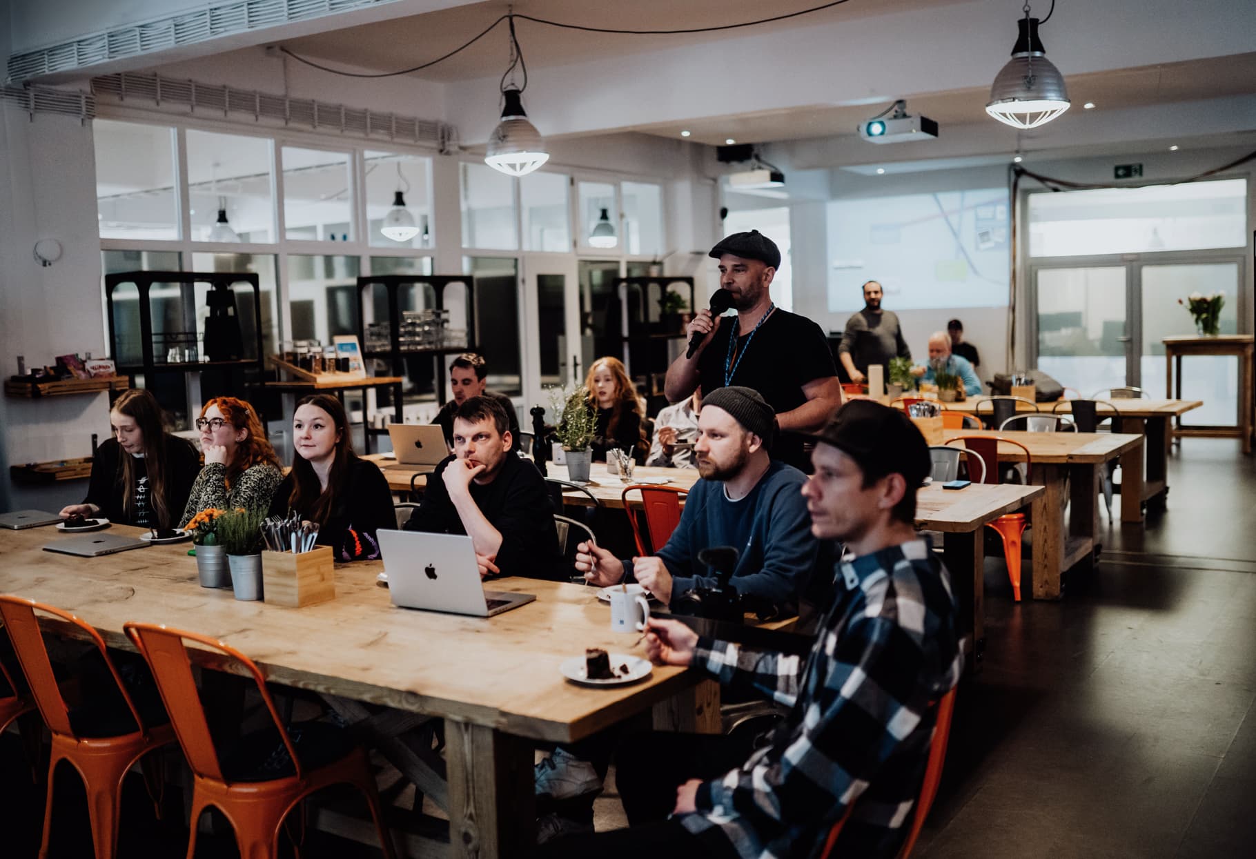 People sitting at large wooden tables with laptops and coffee, attentively following a presentation in a bright, open workspace.